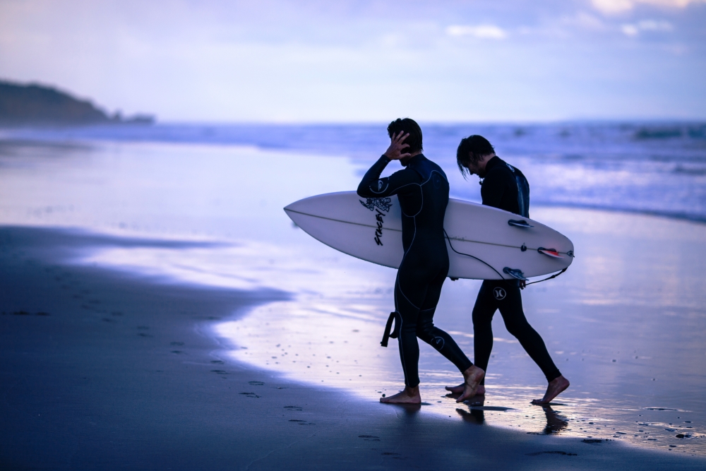 Dwóch mężczyzn na plaży z deską do surfingu Fot. Arnaud Maesureur/Unsplash